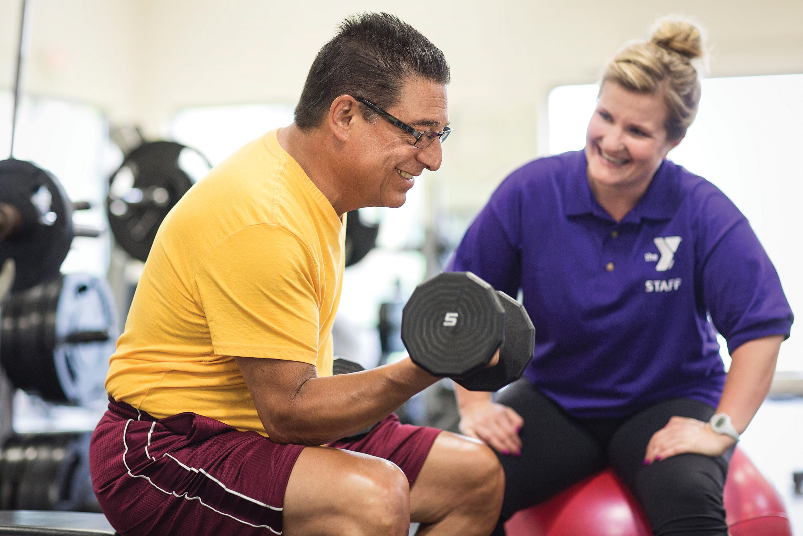 YMCA staff member helping older man lift weights.