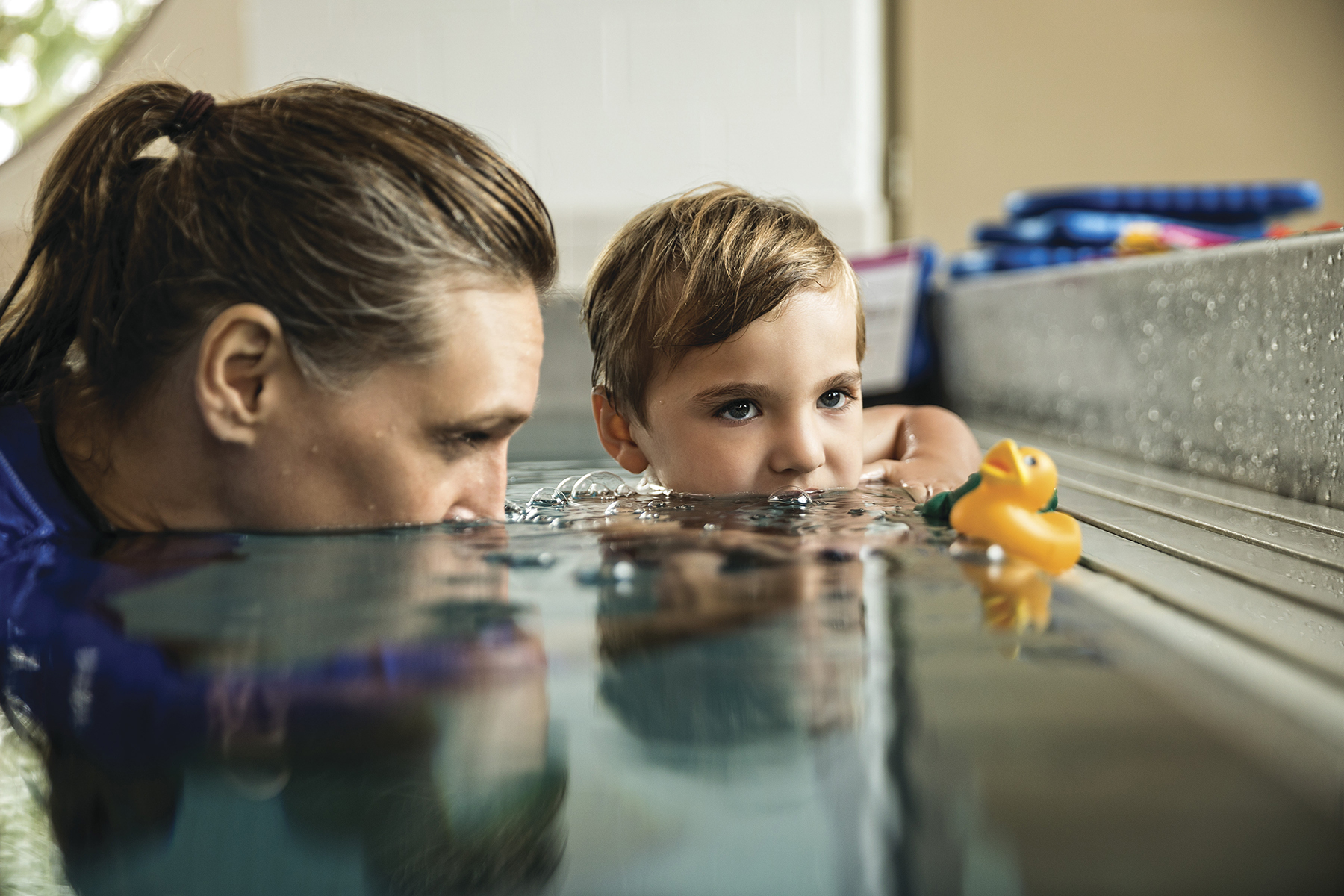 Swim instructor helping young boy swim with toy duck.