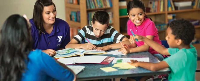 Teacher With Four Young Kids At A Table With Open Books.